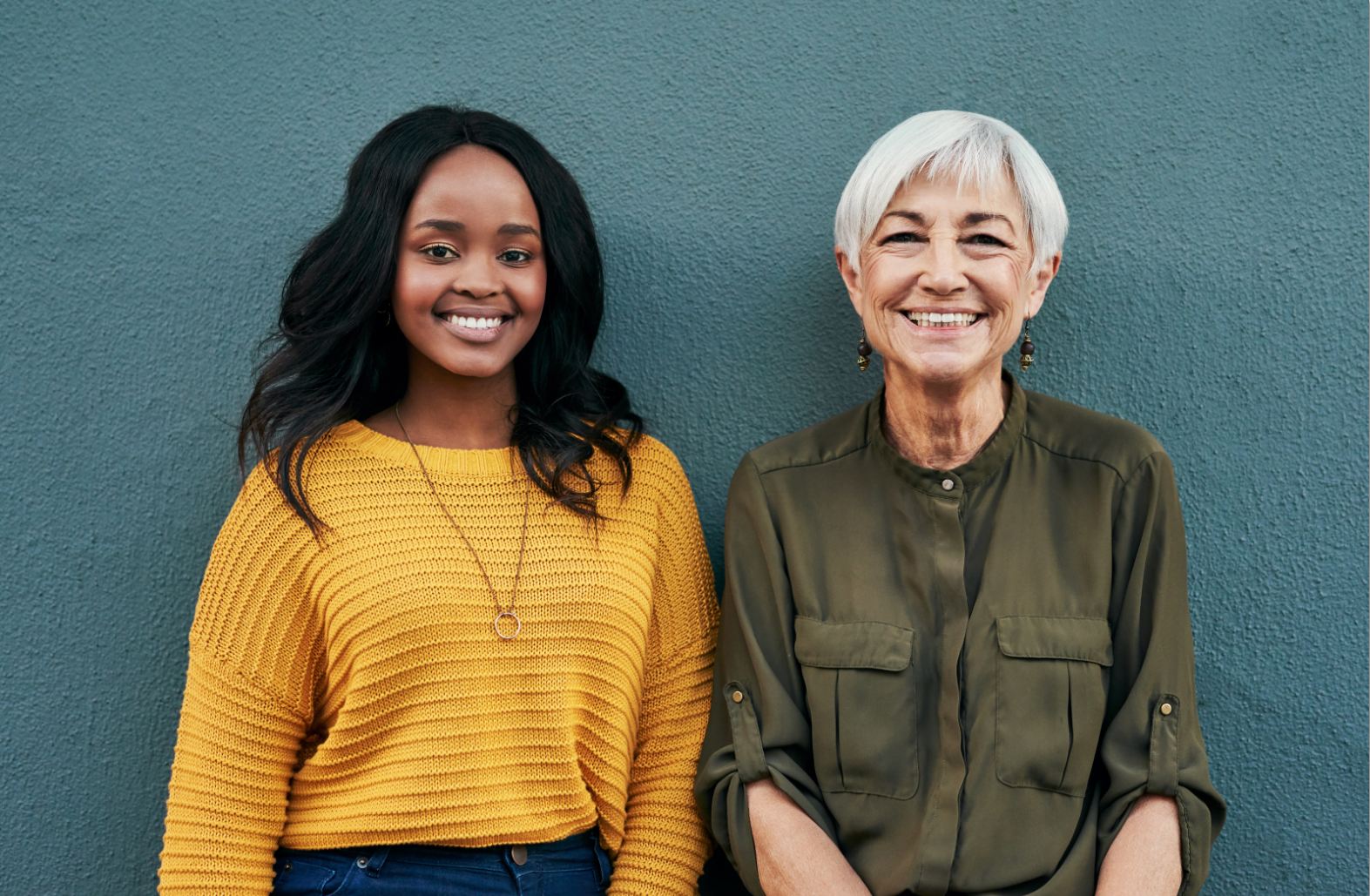 Reach out - young African American woman in a yellow sweater standing next to an older woman with grey hair in an olive green shirt. Both women are smiling and in friendly contact with each other